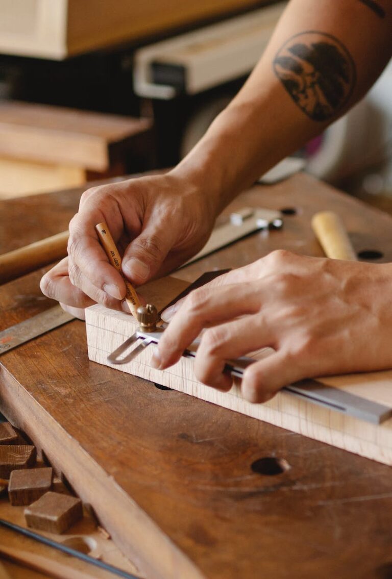 From above of crop unrecognizable tattooed male carpenter measuring wooden plank while creating new object in workshop